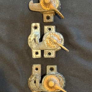 May include: Three antique window latches with ornate metalwork and a gold-colored finish. The latches are in a row, with the top latch having a more simple design and the bottom two latches having more intricate designs.