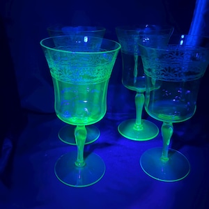 May include: Four vintage green glass goblets with etched floral designs. The goblets have a distinctive glow under a black light, showcasing their unique color and intricate patterns. The stems and bases are also green.