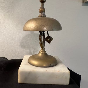Op de afbeelding: Een messingbel met een decoratief handvat en een marmeren voet. De bel wordt gebruikt als een servicebel.