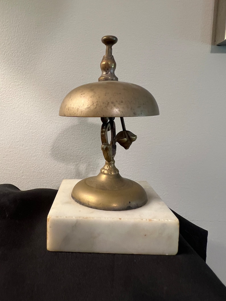 Op de afbeelding: Een messingbel met een decoratief handvat en een marmeren voet. De bel wordt gebruikt als een servicebel.