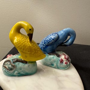 May include: A pair of ceramic figurines of birds perched on a white marble base. The birds are painted in bright colors, one is yellow and the other is blue.