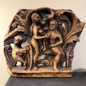 May include: A carved wooden relief sculpture depicting a biblical scene. The artwork features two figures, a woman and a man, with a serpent and a bird. The relief is carved in a warm brown tone, with intricate details in the figures and foliage.