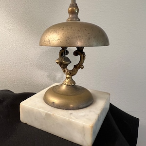 Op de afbeelding: Een messingbel met een decoratief handvat en een marmeren voet. De bel is aangetast en heeft een patina-afwerking.
