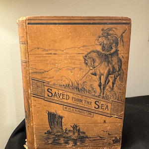 Op de afbeelding: Een vintage boek getiteld "Gered uit de zee" door W.H.G. Kingston. De omslag toont een zwart-witte illustratie van een man te paard die door een landschap met een waterpartij op de achtergrond rijdt. Het boek heeft een bruine leren omslag met zwarte tekst en illustraties.
