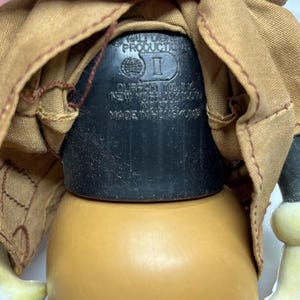 May include: Close-up of a vintage toy doll's base, featuring a black plastic component with embossed text: "WALT DISNEY PRODUCTION", "DURFIN YONG INC. NEW YORK, N.Y. 10019", and "MADE IN HONGKONG". The doll has a tan body and a brown coat.