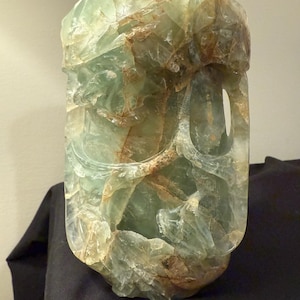 May include: A carved, translucent green and brown fluorite sculpture. The rectangular form has a rounded base and top, with a large, irregular opening on one side. The stone has a rough, textured surface with areas of light and dark green and brown.