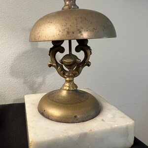 Op de afbeelding: Een messingbel met een decoratieve basis en een witte marmeren standaard. De bel heeft een ronde bovenkant en een sierlijk ontwerp.