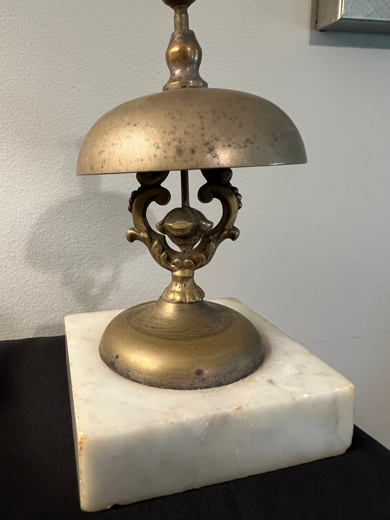 Op de afbeelding: Een messingbel met een decoratieve basis en een witte marmeren standaard. De bel heeft een ronde bovenkant en een sierlijk ontwerp.