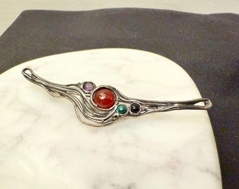 May include: A silver-toned metal hair barrette with an organic, flowing design. It features a large, round, red stone at the center, surrounded by smaller purple, green, and black stones. The barrette is set against a white surface.