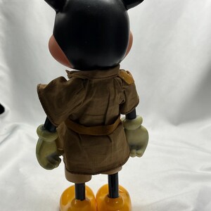 May include: A vintage Mickey Mouse figurine, viewed from the back. The figure has a black head with large ears, a tan face, and yellow gloves and shoes. It wears a brown safari-style outfit with a belt.