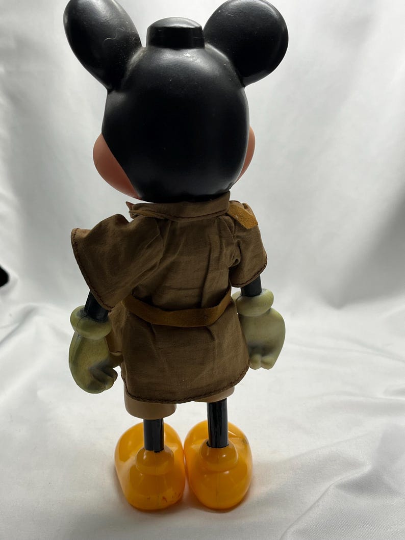 May include: A vintage Mickey Mouse figurine, viewed from the back. The figure has a black head with large ears, a tan face, and yellow gloves and shoes. It wears a brown safari-style outfit with a belt.
