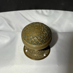 May include: A round brass door knob featuring an intricate embossed pattern of repeating leaf-like shapes and a central geometric square design. The knob is attached to a circular brass mounting plate with visible screw holes at the base.