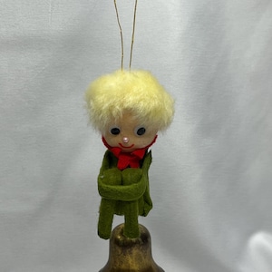 May include: A vintage Christmas ornament featuring a doll-like figure with a yellow fuzzy head, black eyes, and a red bow tie. The figure is dressed in green and stands atop a gold-colored bell. A gold string is attached for hanging.