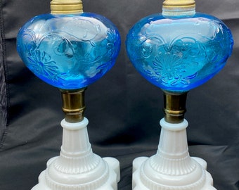 May include: Two antique oil lamps. Each lamp features a heart-shaped, turquoise glass reservoir with floral detailing. The reservoirs sit atop brass stems and white, star-shaped bases. The lamps have brass tops.