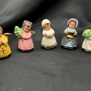 May include: Set of five small, hand-painted porcelain figurines of angels. Each angel is wearing a different colored dress and has a unique accessory, such as a tree, a book, or a bouquet of flowers.