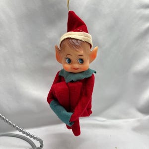 May include: A vintage Christmas elf ornament. The elf has a red felt outfit with green cuffs and collar, a red pointed hat with a cream-colored band, and a painted face with blue eyes. A gold string is attached for hanging.