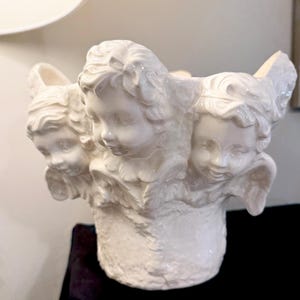 May include: A white ceramic vase featuring three cherubic faces with wings. The faces are detailed with curly hair and gentle expressions. The vase has a textured base and is likely used for decorative purposes.