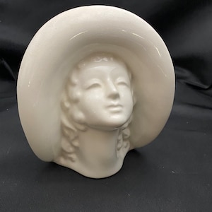 May include: White ceramic sculpture of a woman's head wearing a wide-brimmed hat. The sculpture is a simple, minimalist design.