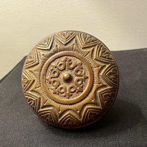 May include: A brass door knob with an intricate, ornate design. The knob features a central circle with a raised, circular button in the center. The circle is surrounded by a ring of smaller, decorative circles. The outer edge of the knob is decorated with a series of repeating, geometric patterns.