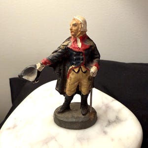 May include: A painted metal figurine of a historical figure wearing a dark blue coat with red accents, tan breeches, and black boots. The figure holds a black hat in an outstretched right hand and stands on a circular base, facing to the left.
