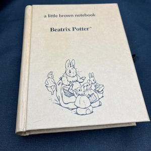 May include: A brown notebook with a black and white illustration of Peter Rabbit and his sisters on the cover. The text "a little brown notebook" and "Beatrix Potter" is printed on the cover.