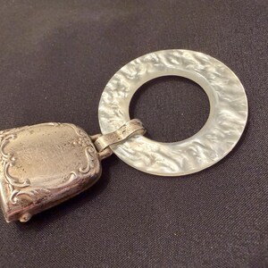 May include: An antique silver-toned rattle with a rectangular bell-shaped section and a round, pearlescent ring. The bell features ornate floral detailing. The ring has a marbled appearance. The rattle is connected by a small metal band.
