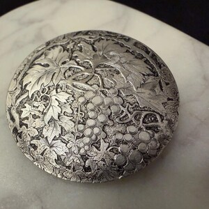May include: A round, silver-toned decorative piece with a detailed relief of grape vines and leaves. The design is centered on the object, with the grapes and foliage filling the surface. The piece has a vintage aesthetic.