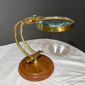 May include: A brass magnifying glass mounted on a curved, adjustable brass arm attached to a circular, polished wooden base. The magnifying lens is encased in a brass rim, featuring a small adjustment screw at the top of the curved support arm.
