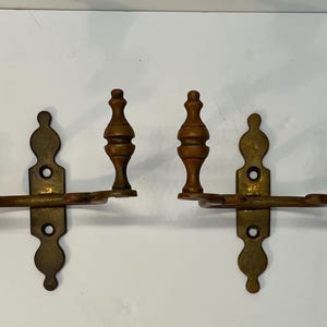 May include: Two antique brass wall sconces with wooden accents. Each sconce features a decorative backplate, a horizontal arm, and three wooden finials. The sconces are designed for mounting on a wall and add a vintage touch.