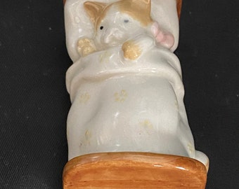May include: A ceramic figurine of a cat sleeping in bed. The cat is white with brown and orange markings. The bed is brown with a white blanket and yellow flowers.