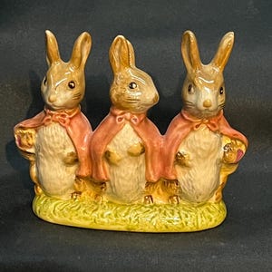 May include: A ceramic figurine featuring three brown rabbits standing side-by-side on a green grassy base. Each rabbit wears a pink cape tied at the neck with a bow. The rabbits have white chests, pointed ears, and detailed facial features.