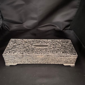 May include: A silver rectangular box with an ornate floral design. The box has a hinged lid and sits on four small feet.