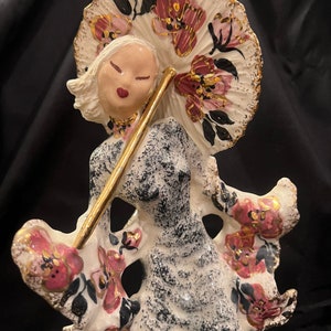 May include: A white ceramic figurine of a woman holding a gold and pink umbrella. The woman is wearing a black and white dress with pink flowers. The figurine is in a vintage style.