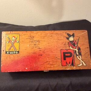 May include: Vintage wooden box with the text "x-acto" and a graphic of a person holding two crossed knives. The box has a red painted section and a cartoon figure leaning on a red "PM" sign.
