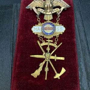 May include: A gold-toned metal pin with a spread-winged eagle, a chain with a circular emblem, and a cross with five weapons. The eagle has a banner that reads "F.F. & C.". The circular emblem has a red, white, and blue design and reads "Improved Order of Red Men, Tote.". The pin is on a red velvet background.