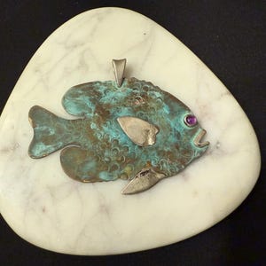 May include: A turquoise and bronze fish pendant with a purple gemstone eye, set on a white marble-like stone. The fish has silver-colored fins and is mounted on a white, oval-shaped stone. The background is black.