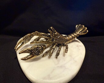 May include: A detailed bronze-colored metal crawfish figurine with textured claws, legs, and tail, resting on a white, heart-shaped marble base. The crawfish is a decorative item, possibly a paperweight or ornament.