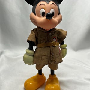 May include: Vintage Mickey Mouse figurine. The figure has a black head and ears, a tan face, and a brown outfit with a belt. It wears yellow shoes and green gloves. The figurine is a collectible item.