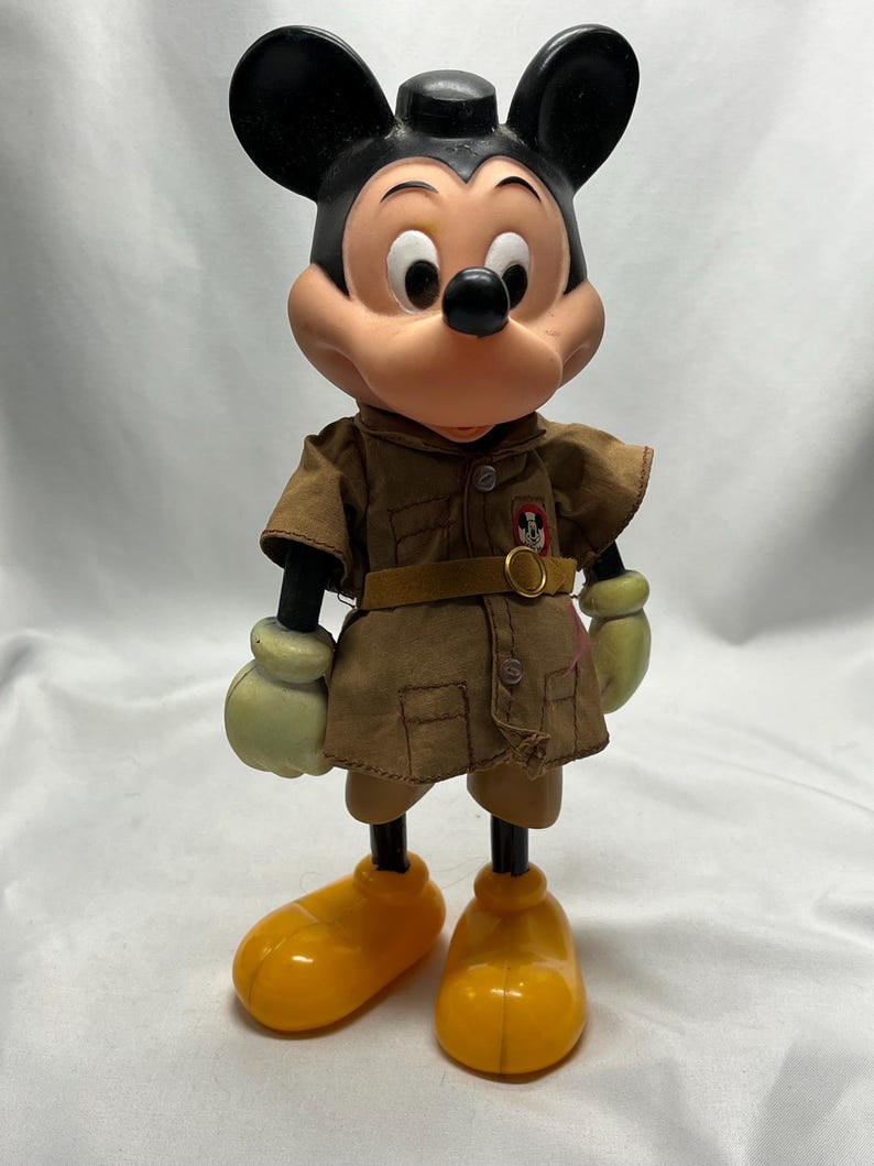 May include: Vintage Mickey Mouse figurine. The figure has a black head and ears, a tan face, and a brown outfit with a belt. It wears yellow shoes and green gloves. The figurine is a collectible item.