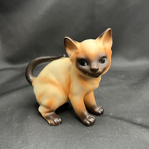 Op de afbeelding: Een keramieken Siamese kattenfiguur met bruine en beige markeringen, blauwe ogen en een gekrulde staart. De kat zit. Het beeldje heeft een glanzende afwerking en staat tegen een donkere achtergrond.