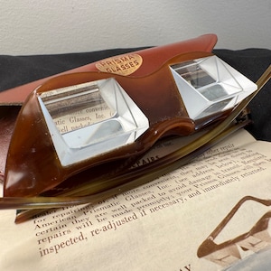 May include: A pair of brown Prisma Glasses in a brown leather case. The glasses are designed to magnify images and have a clear plastic lens. The case has the words "Prisma Glasses" printed on it.