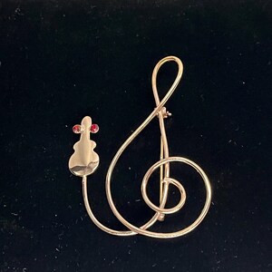 May include: A gold-toned brooch featuring a treble clef design with a small violin and two red gemstones. The brooch is set against a black background, highlighting its elegant and musical theme.