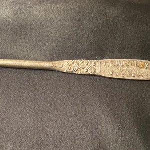 May include: A vintage metal crochet hook with a curved end and a decorative handle. The handle is embossed with floral designs and the text "E. MOSELEY CO. BOSTON". The hook is silver in color.