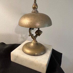 Op de afbeelding: Een messingbel met een marmeren voet. De bel heeft een decoratief handvat en een gepatineerde afwerking.
