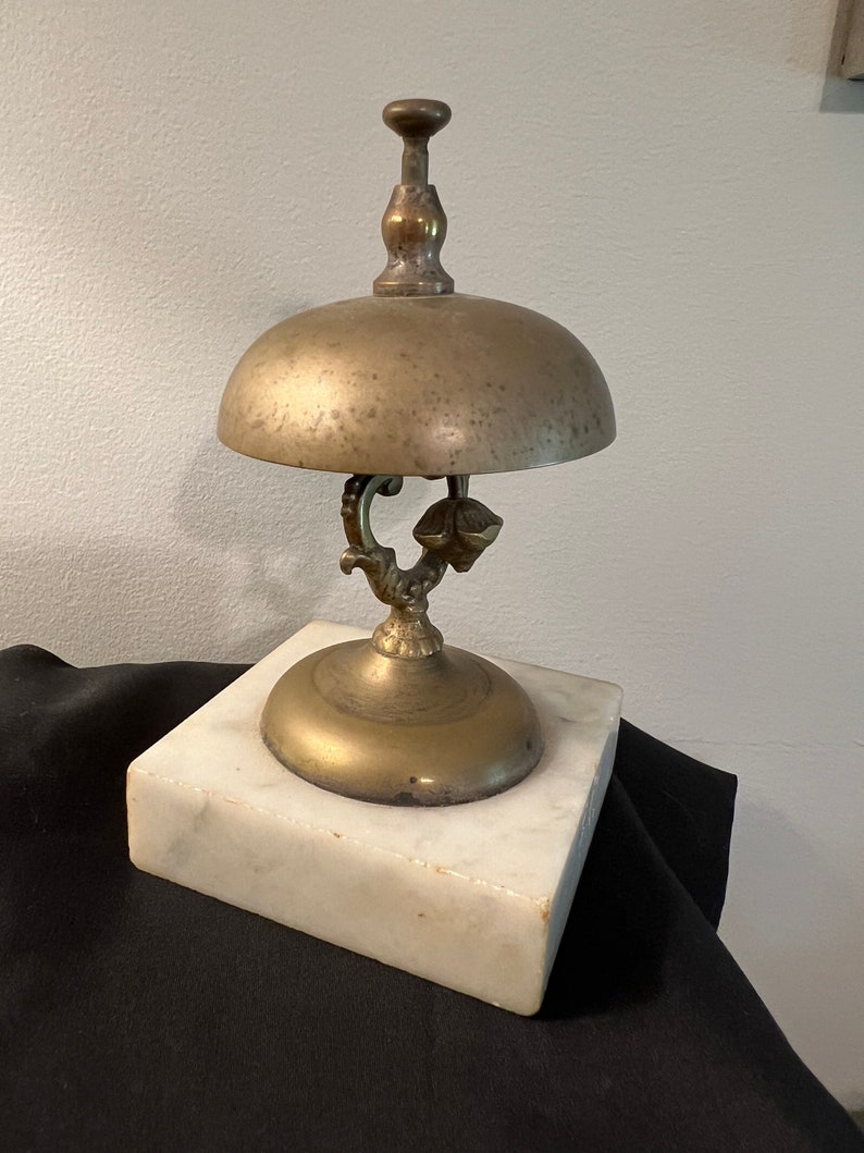 Op de afbeelding: Een messingbel met een marmeren voet. De bel heeft een decoratief handvat en een gepatineerde afwerking.