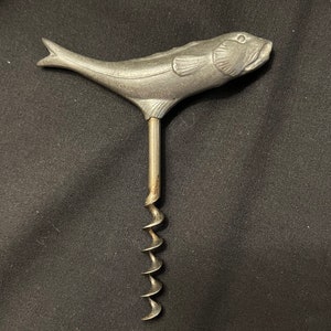May include: A silver metal fish-shaped corkscrew with a detailed design. The corkscrew has a long, thin handle and a spiral corkscrew.