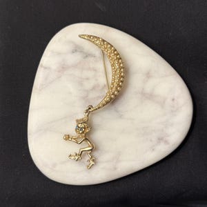 May include: A gold-toned brooch featuring a crescent moon adorned with small, round beads. A whimsical figure with blue eyes and a hat hangs from the moon. The brooch is set on a white, oval-shaped stone.