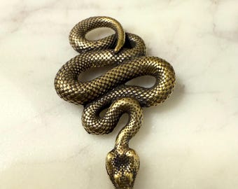 May include: A detailed, antique-style brass snake figurine. The snake is coiled with its head raised, showcasing textured scales and a detailed head. The figurine is set against a white and gray marble surface.