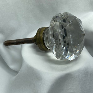 Puede incluir: Un pomo de puerta de cristal transparente con un diseño facetado y una base de latón. El pomo está unido a una varilla de metal.