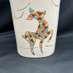May include: White ceramic vase with a stylized deer design. The deer is painted in shades of pink, blue, yellow, and brown. The vase has a speckled glaze and gold accents.